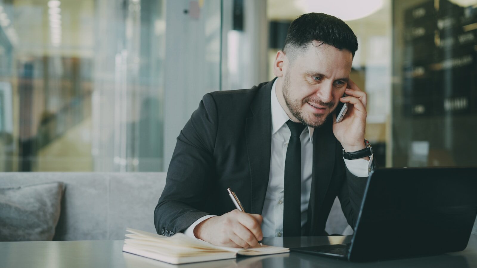 Man in suit talking on phone while writing in notebook.